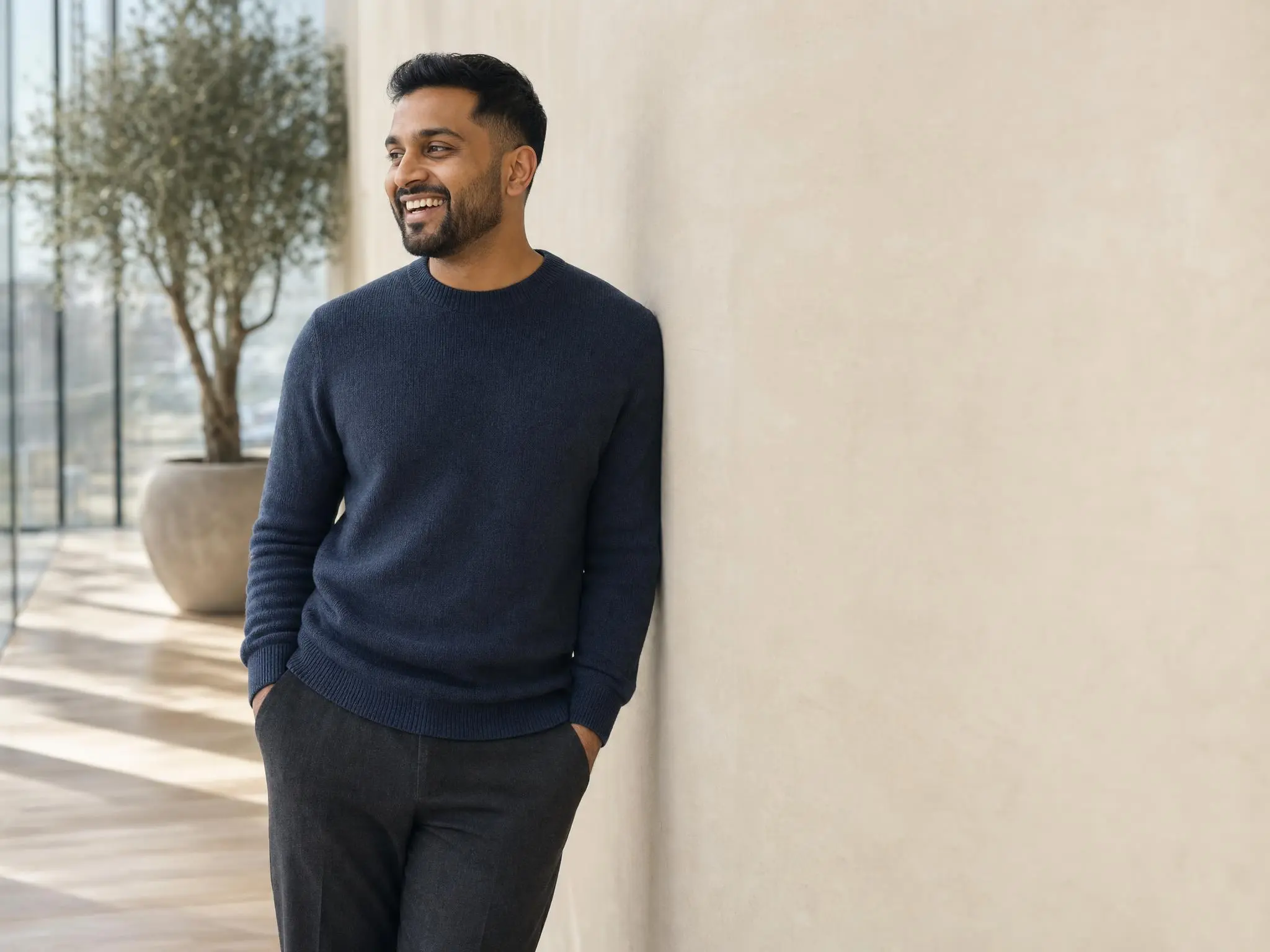 A man in a navy sweater leaning against a sunlit atrium wall, hands in his pockets, laughing at something out of frame.