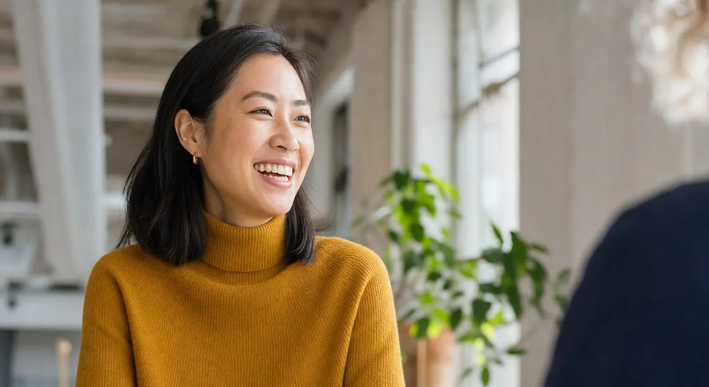 A woman in a mustard roll-neck laughing mid-conversation in a bright, plant-filled office.